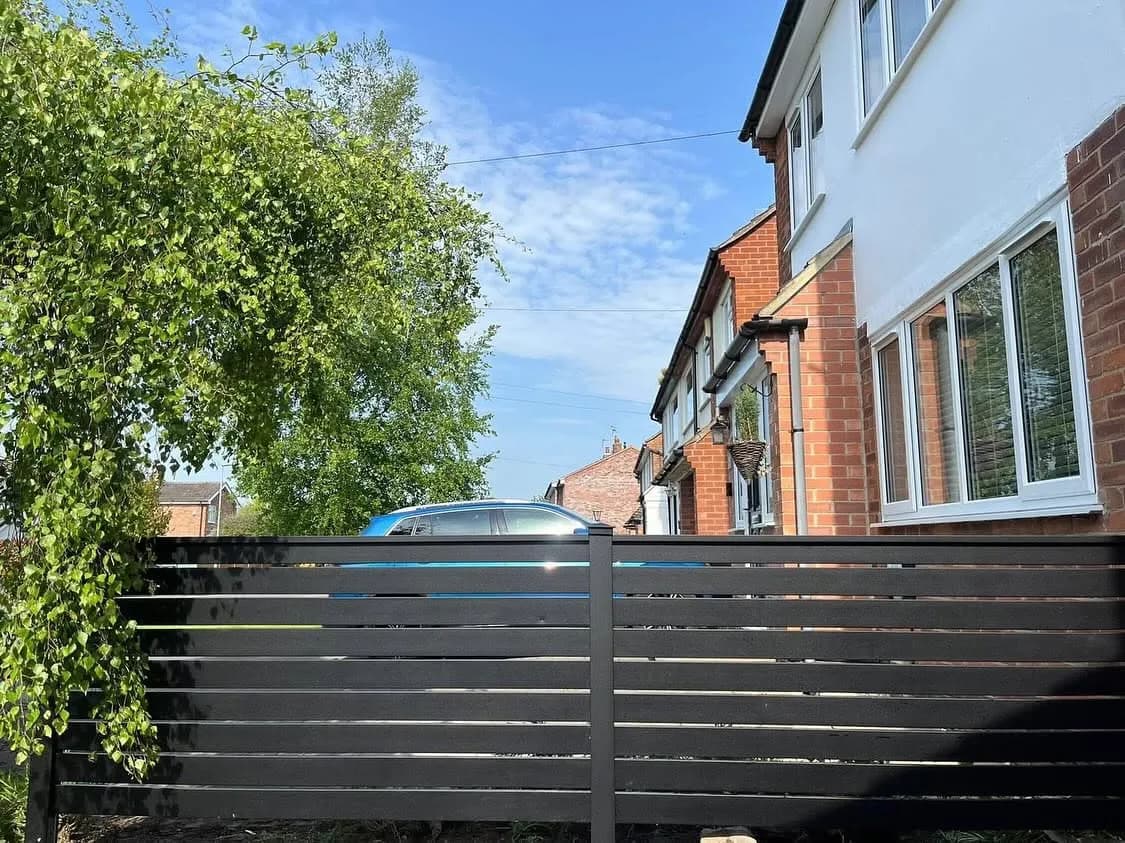 Anthracite slatted aluminium fence beside rendered and brick houses with a climbing plant