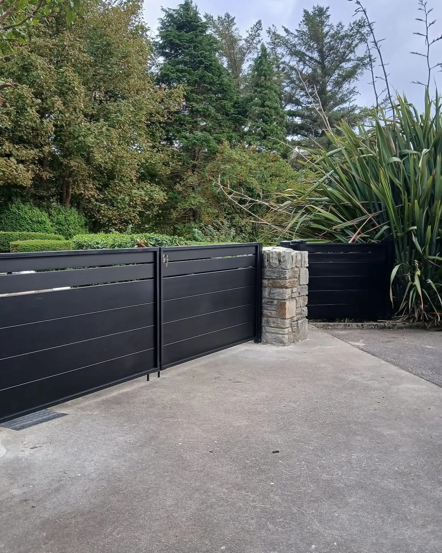 Anthracite aluminium driveway gate beside a stone pillar and coastal planting